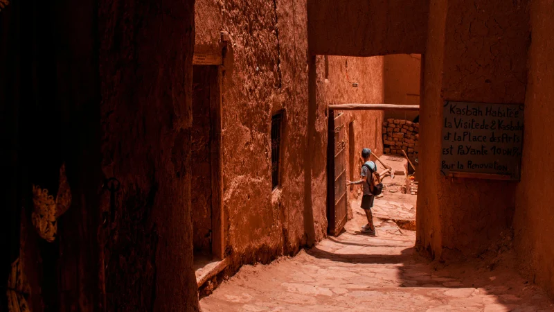 Traveler walking through a narrow, sunlit alley of a traditional Moroccan kasbah with reddish clay walls and rustic wooden doors.