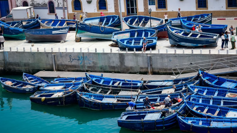 Colorful fishing boats moored in a harbor, with some individuals on the shore and in the boats, surrounded by blue water and a backdrop of an old stone building.