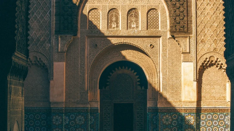 Ornate courtyard with intricate tilework, arched entrances, detailed carvings, and a rectangular reflecting pool lit by sunlight.