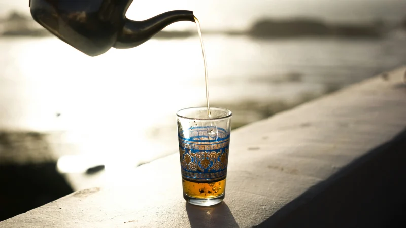 A hand pours tea from a metal teapot into a decorated glass on a sunlit outdoor ledge, with a blurred body of water in the background.